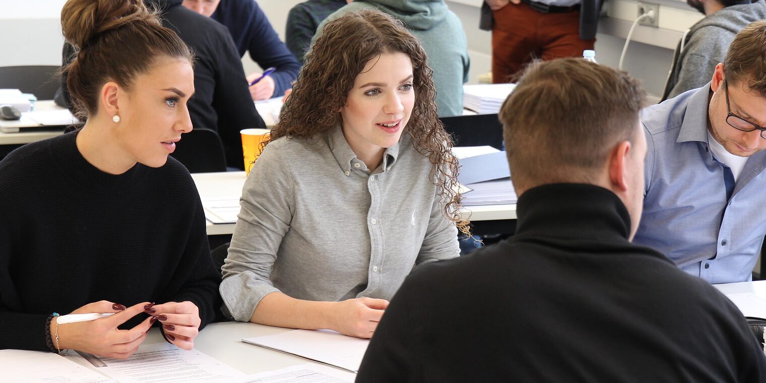 Symbolbild für Weiterbildung, 5 junge Menschen sitzen zusammen an einem Tisch in einem Klassenzimmer