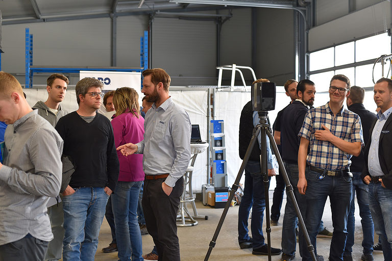 3D-Laser-Aufmaß, das Messgerät steht auf einem Stativ in einer Halle und wird von Besuchenden angeguckt