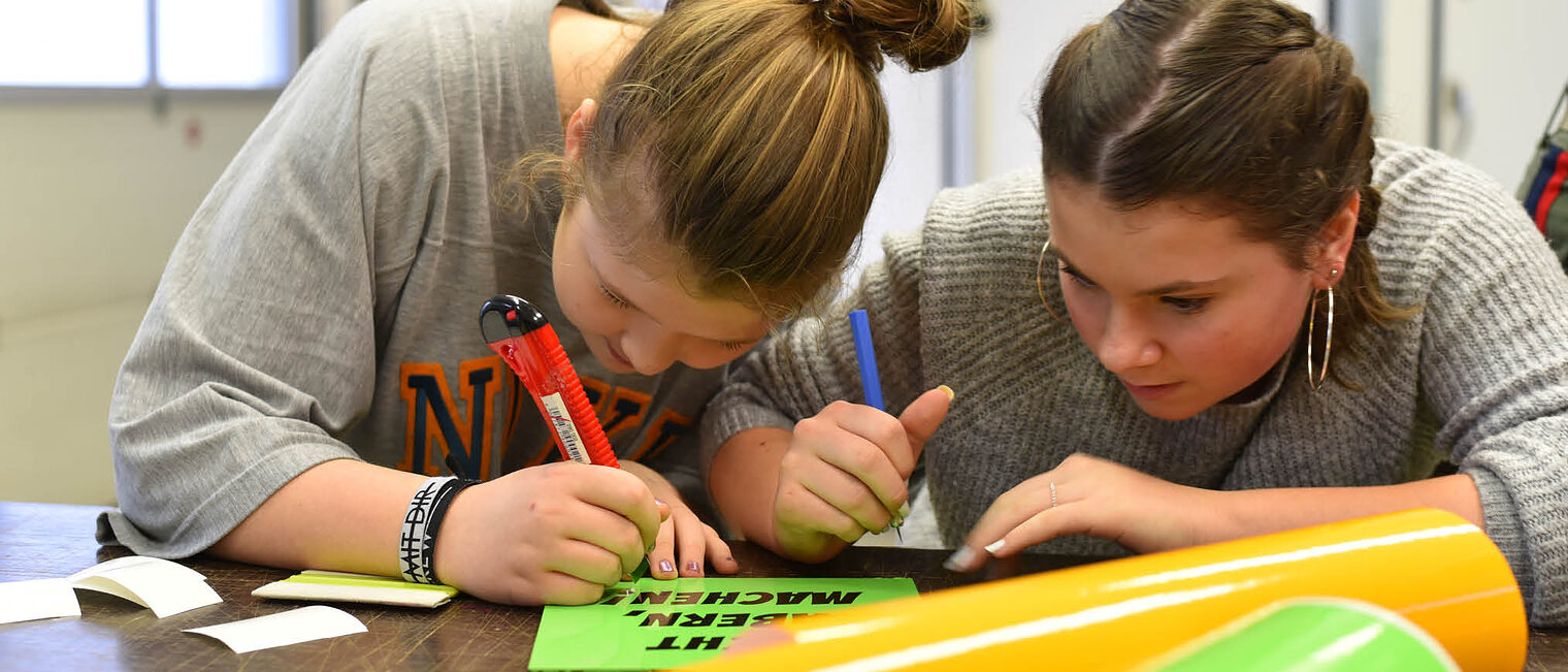 Zwei Mädchen arbeiten an Schriftzug mit Cutter und Stift