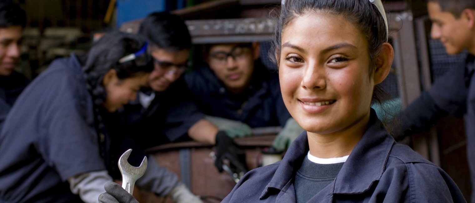 Eine junge Nepalesin im Blaumann steht vor Gruppe junger Leute, die ein Auto reparieren