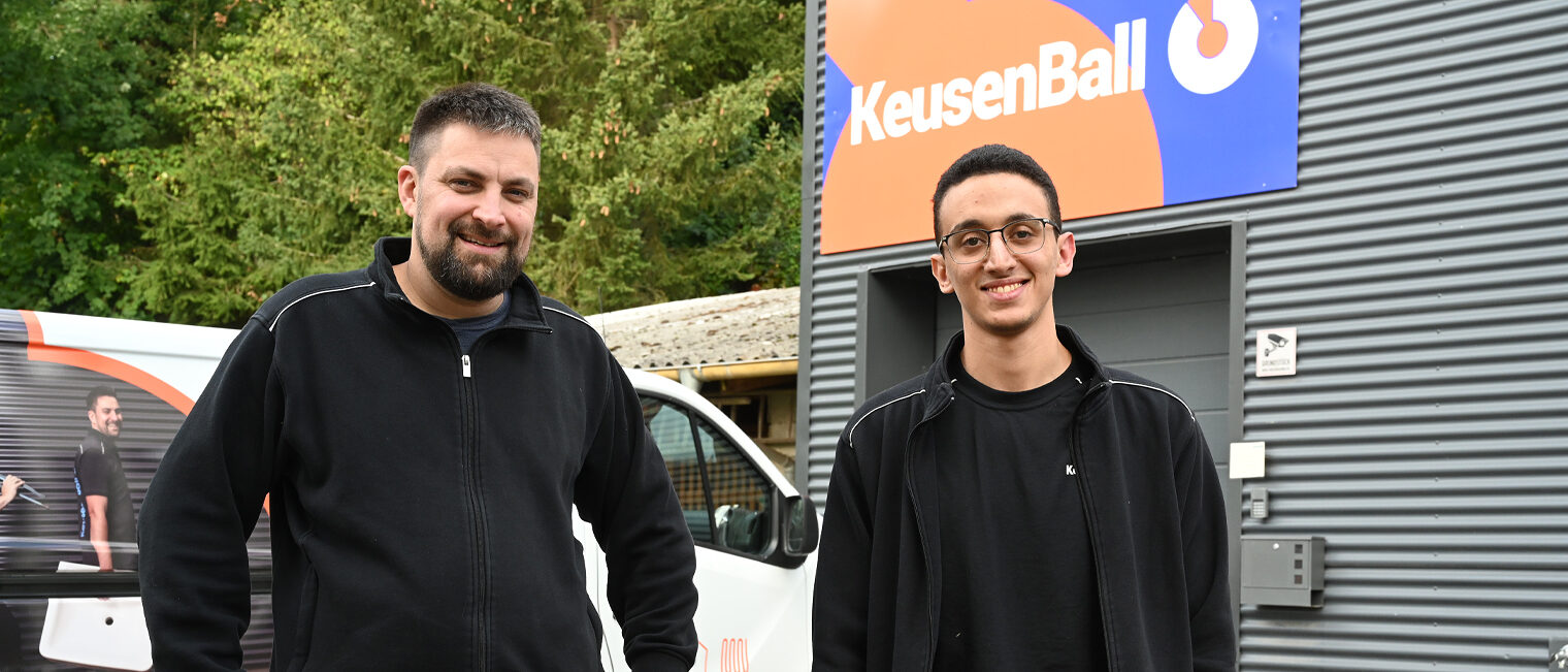 Betriebsinhaber und sein Auszubildender stehen auf dem Hof des Betriebes, beide schauen in Kamera