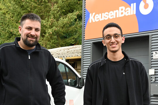 Betriebsinhaber und sein Auszubildender stehen auf dem Hof des Betriebes, beide schauen in Kamera