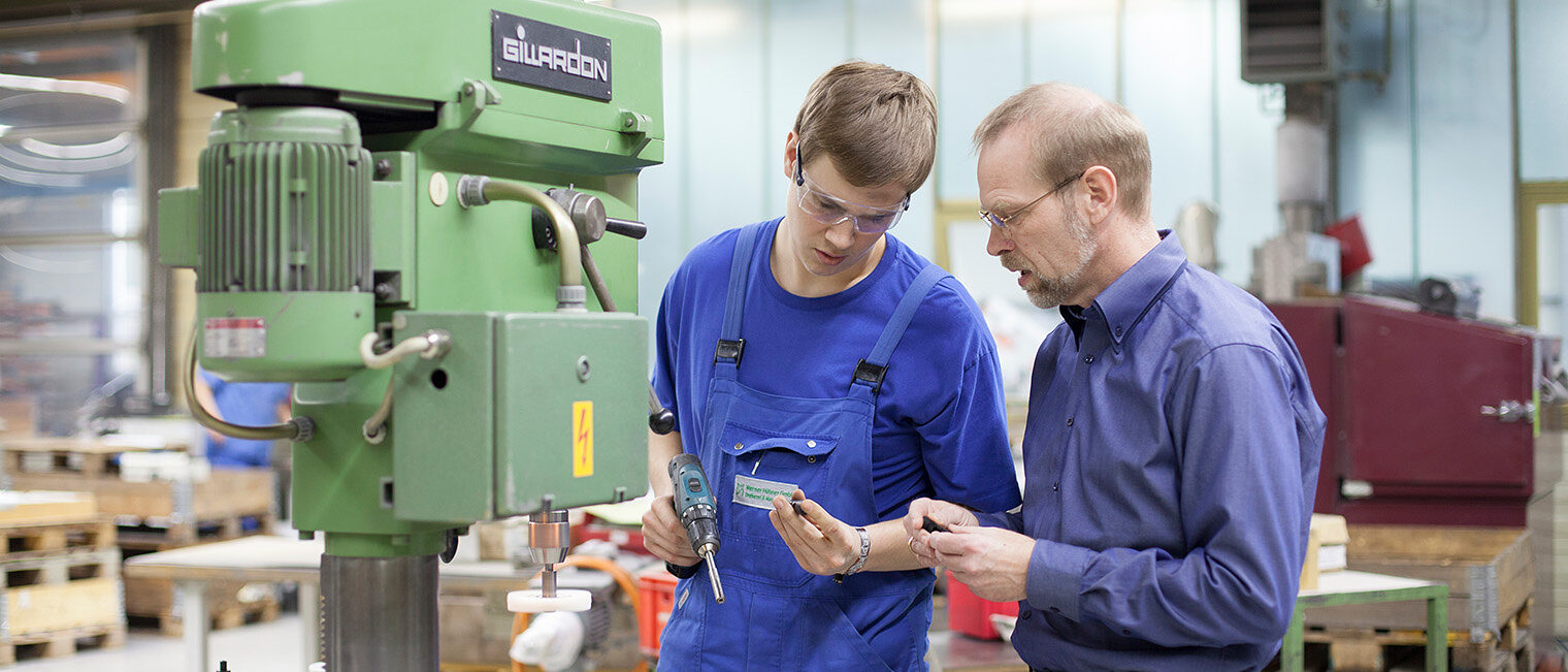 Ein Auszubildender mit seinem Meister, beide stehen in einer Werkstatt vor einer Maschine, beide tragen Schutzbrillen