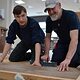 Peter Meeks und Auszubildender Dominik Bund knien auf dem Boden und zeigen das Verlegen von Klick-Vinyl 