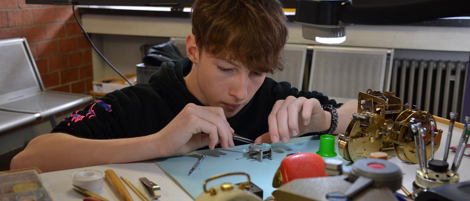 Schüler arbeitet an Uhrmechanik mit Pinzette an Tisch mit Werkzeugen