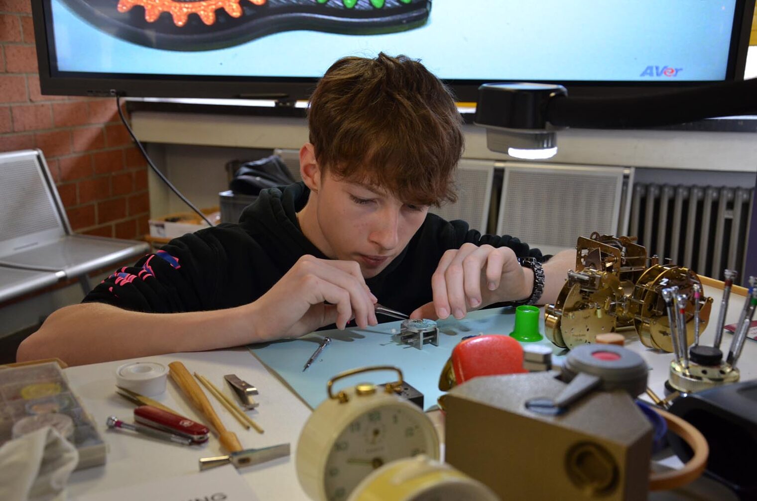 Schüler arbeitet an Uhrmechanik mit Pinzette an Tisch mit Werkzeugen