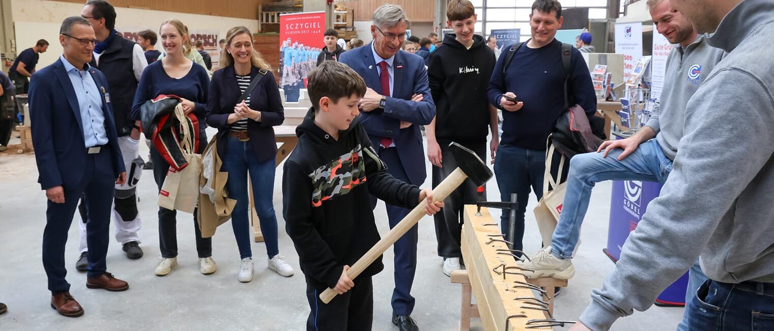 Besucher des Macher-Tags schlägt langen Nagel mit großem Hammer in Holz-Balken vor Publikum, das im Kreis steht.