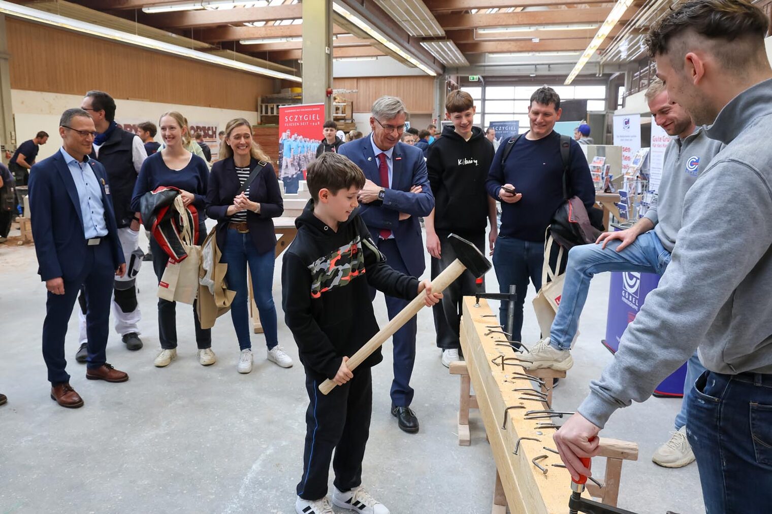 Besucher des Macher-Tags schlägt langen Nagel mit großem Hammer in Holz-Balken vor Publikum, das im Kreis steht.