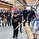 Besucher des Macher-Tags schlägt langen Nagel mit großem Hammer in Holz-Balken vor Publikum, das im Kreis steht.