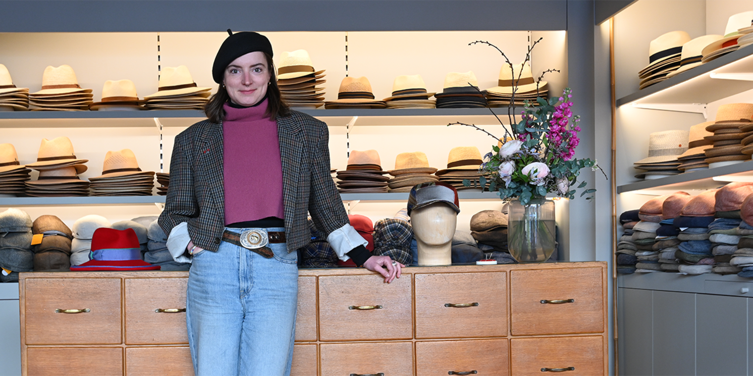 Modistin Laura Zieger in ihrem Geschäft in Würzburg vor einer Kommode, auf der Hüte liegen; im Hintergrund Regale mit Hüten.