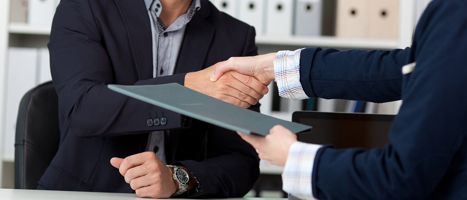 Zwei Personen mit Hemd und Anzug sitzen an Schreibtisch und reichen sich die Hand und übergeben Unterlagen