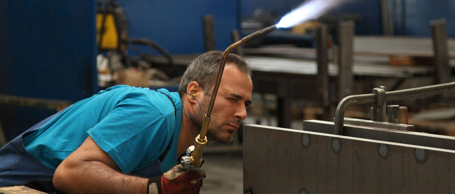 Metallbauer mit Schweißgerät in der Hand inspiziert die Schweißnaht Metallbauer in Schutzkleidung mit Schweißgerät in der rechten Hand inspiziert gebückt die Schweißnaht