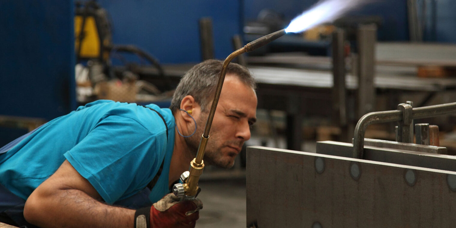 Metallbauer in Schutzkleidung mit Schweißgerät in der rechten Hand inspiziert gebückt die Schweißnaht 