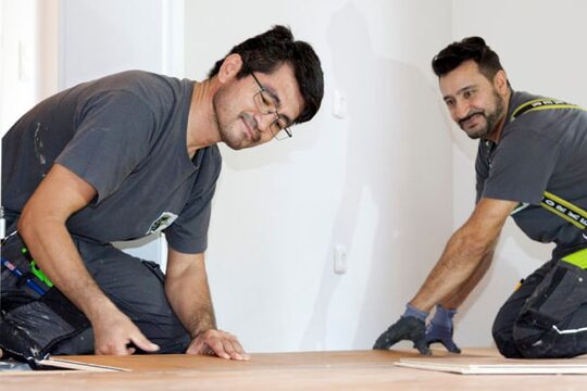 Zwei Männer knien in Arbeitskleidung auf dem Boden und verlegen Parkett