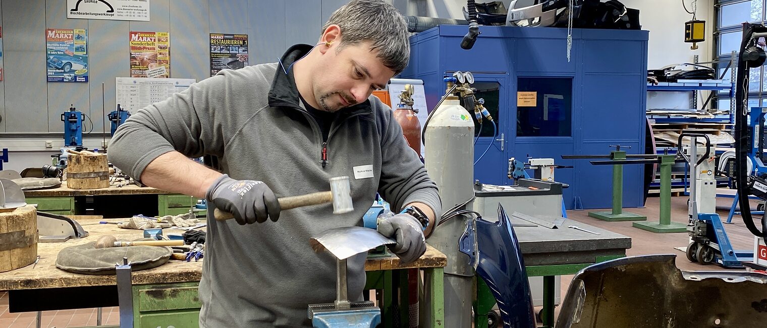 Blechbearbeitung Ein Mann bearbeitet ein eingespanntes Metallblech mit Hammer, trägt Handschuhe, im Hintergrund Werkzeuge und Gasflaschen.