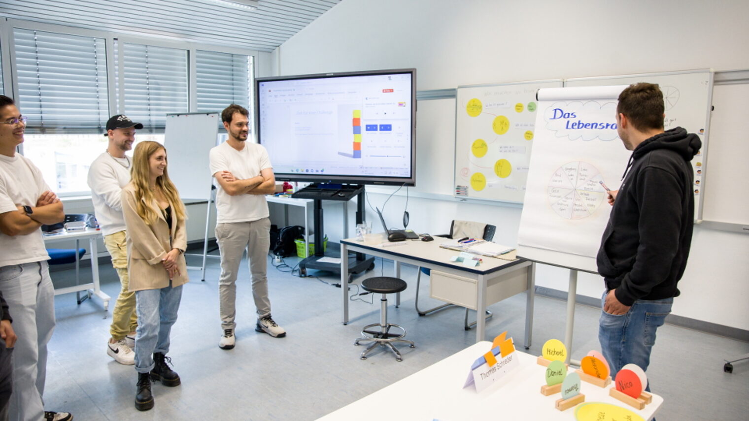 Eine Gruppe junger Menschen steht im Seminarraum, ein Mitschüler erklärt am Flipchart "Das Lebensrad“.