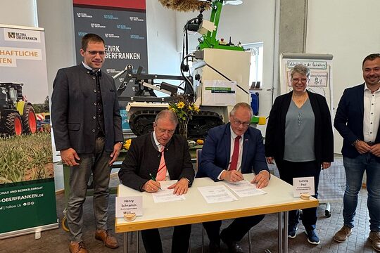 Fünf Personen sind auf dem Bild. Zwei sitzen am Tisch und unterschreiben einen Vertrag. Drei Personen stehendrumherum 