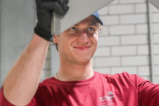 Junger Mann in rotem Oberteil (WorldSkills Logo) hält mit Arbeitshandschuhen ein Blech in die Höhe