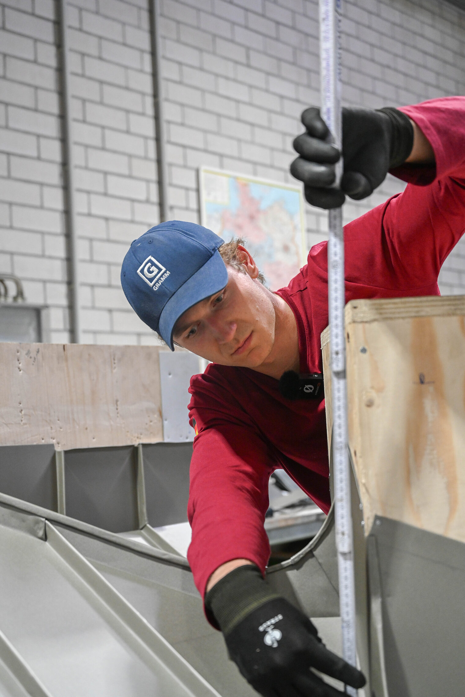 Junger Mann in rotem Longsleeve mit Arbeitshandschuhen misst Holzmodell mit Meterstab ab