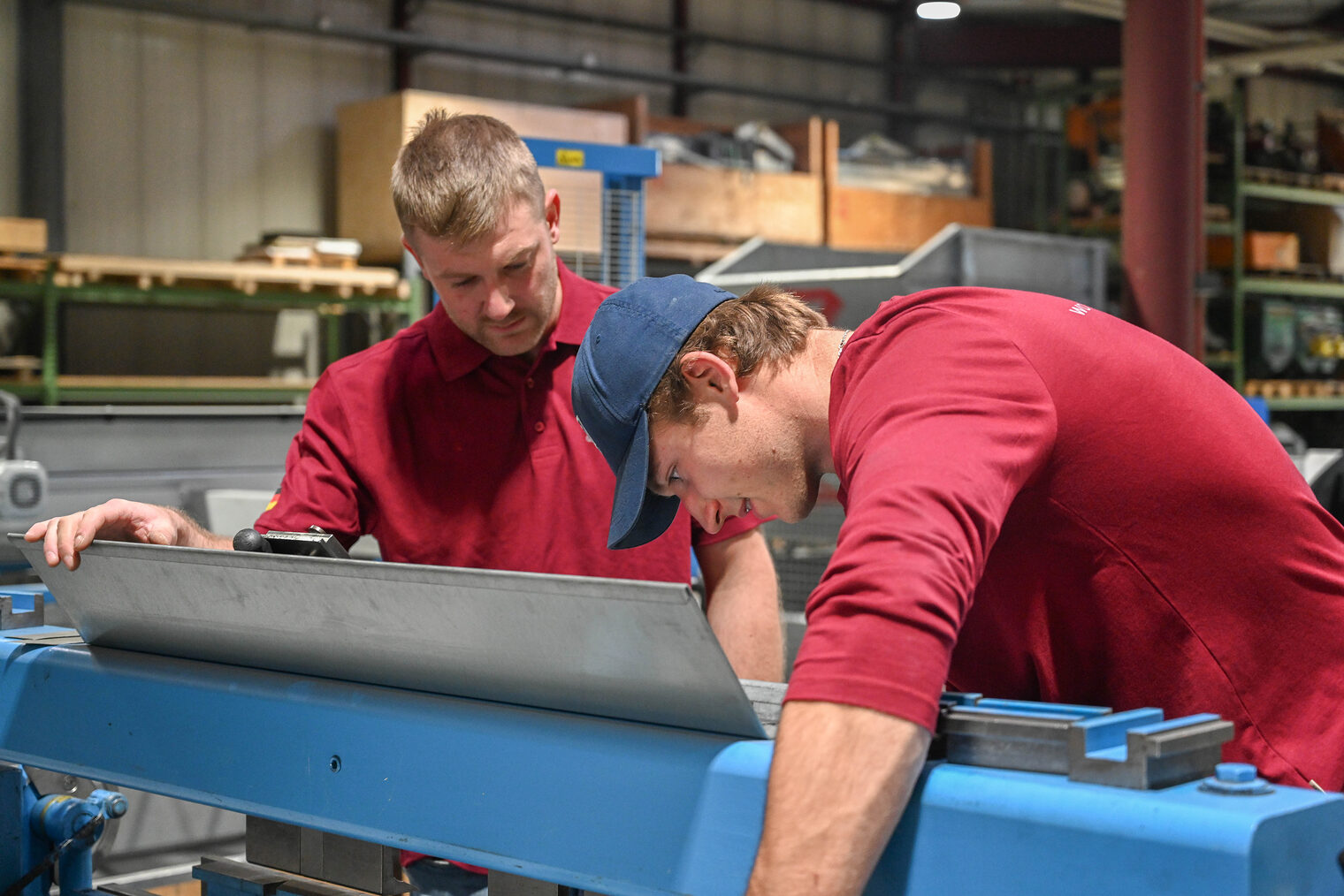 Junger Mann in rotem Longsleeve mit Arbeitshandschuhen arbeitet an Maschine zum Blechbiegen. Trainer steht daneben