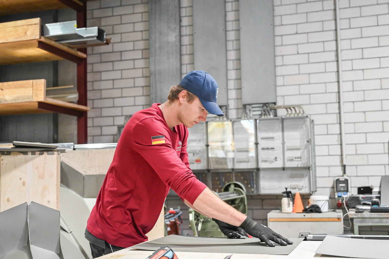 Junger Mann in rotem Longsleeve und blauer Cap arbeitet mit Arbeitshandschuhen an einem Blech