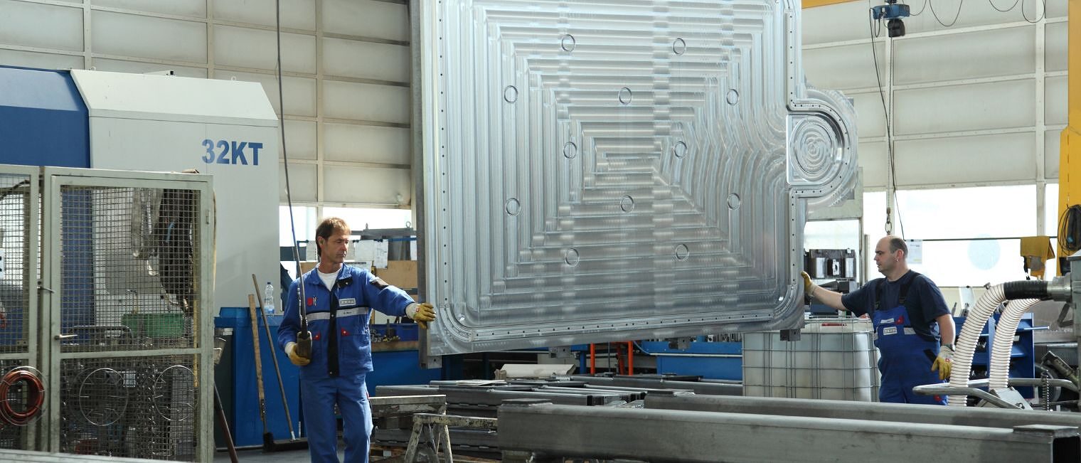 Zwei Männer in Blaumännern und Arbeitshandschuhen stehen an einer riesigen Metallplatte, die im Raum zwischen ihnen hängt