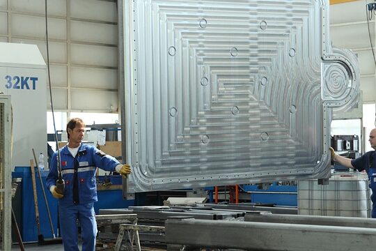 Zwei Männer in Blaumännern und Arbeitshandschuhen stehen an einer riesigen Metallplatte, die im Raum zwischen ihnen hängt