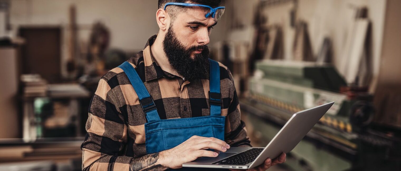 Mann mit kariertem Hemd und blauer Latzhose steht in einer Werkstatt und schaut auf einen Laptop, den er in den Händen hält