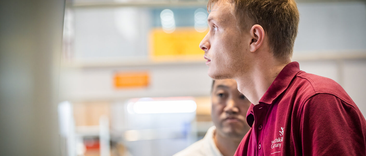 Junger Mann im Seitenprofil, Polohemd dunkelrot mit Aufschrift WorldSkills Germany, Lichtschein von vorne in sein Gesicht
