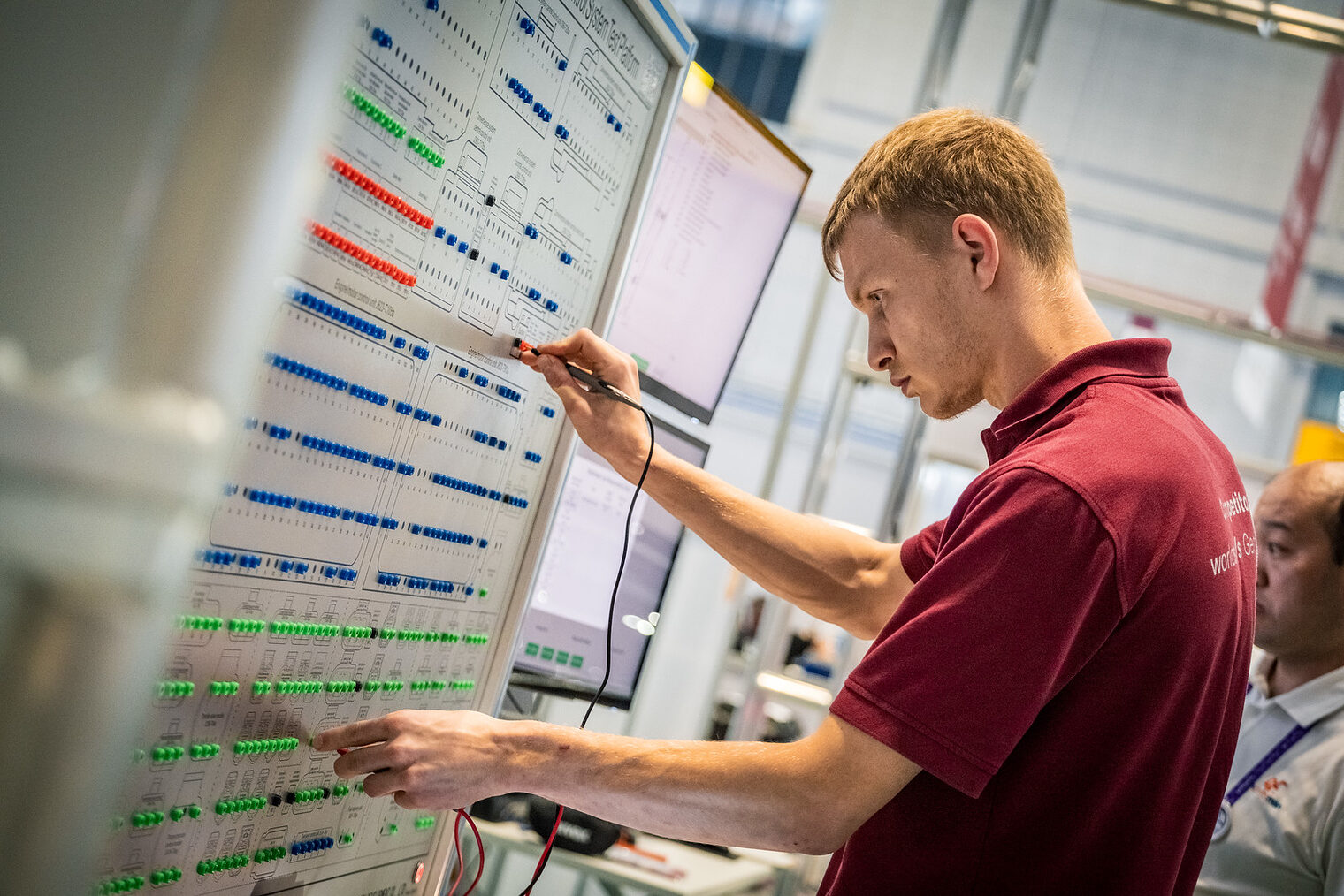 Junger Mann an einem technischen Board mit Testgerät in der Hand, rotes T-Shirt, Foto von der Seite