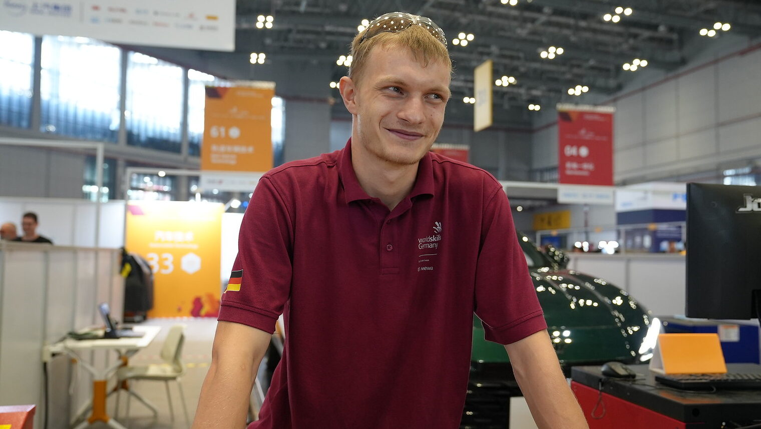 Junger Mann mit rotem Polohemd, Aufschrift: Worldskills Germany, Deutschlandflagge auf dem Ärmel, Lächelt