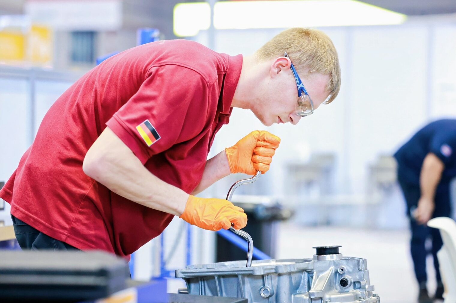 Junger Mann mit Schutzbrille und Handschuhen über ein Autoteil gebeugt, Polohemd mit Deutschlandflagge auf dem Ärmel
